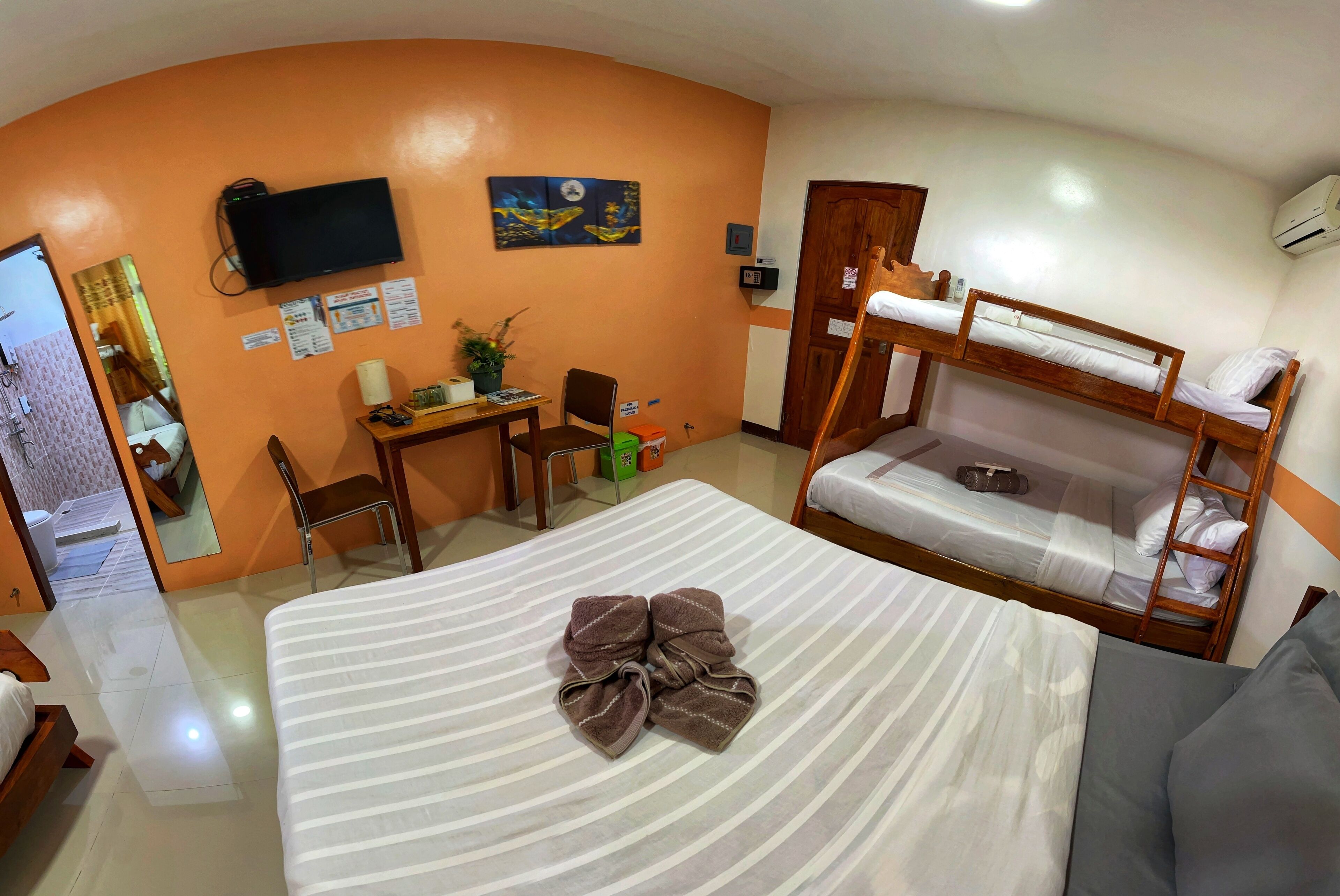 A hotel room with a bunk bed, a desk with a chair, a television mounted on the wall, and a bed with a striped blanket and a brown towel.