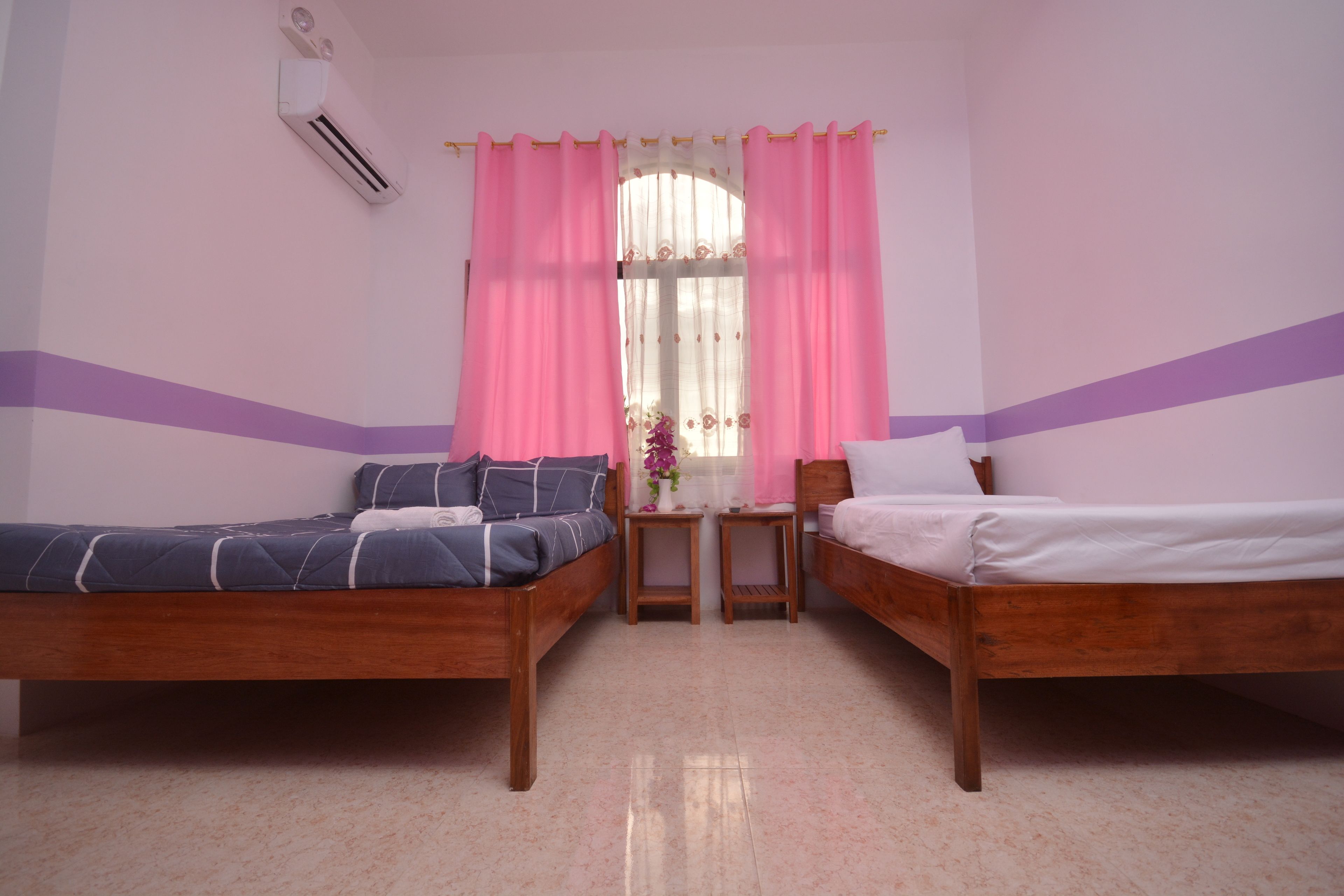 A room with two wooden beds, a small table, and a window with pink curtains.