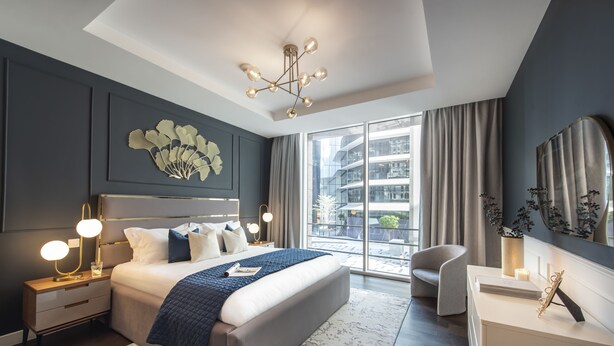 A modern bedroom with a large bed, bedside lamps, a nightstand, a chair, a desk, and a large window with curtains.