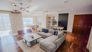 A spacious living room with a sectional sofa, a coffee table, and a dining area with chairs. There is a large window providing a view of the city.
