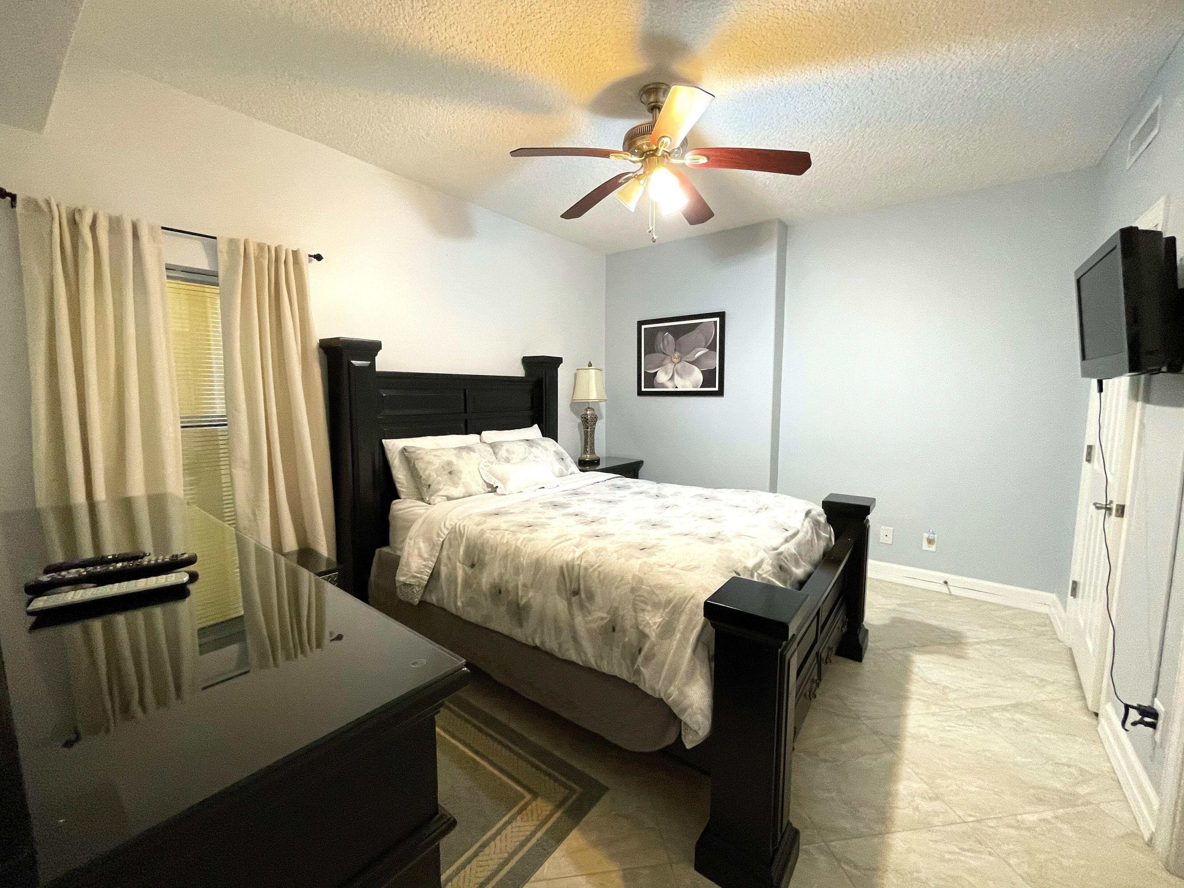 A bedroom with a bed, bedside table, lamp, ceiling fan, and a TV mounted on the wall.