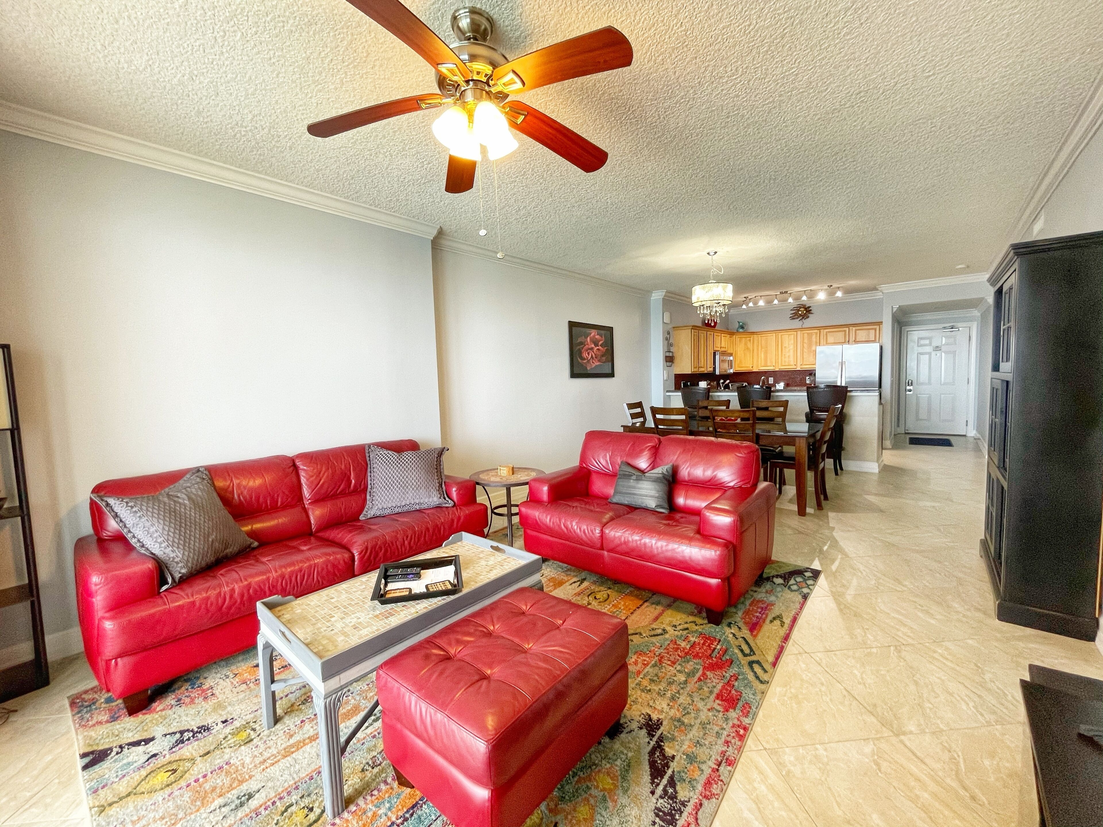 A living room with a red leather sofa, a matching ottoman, and a coffee table. There is a ceiling fan, a chandelier, and a kitchen in the background.