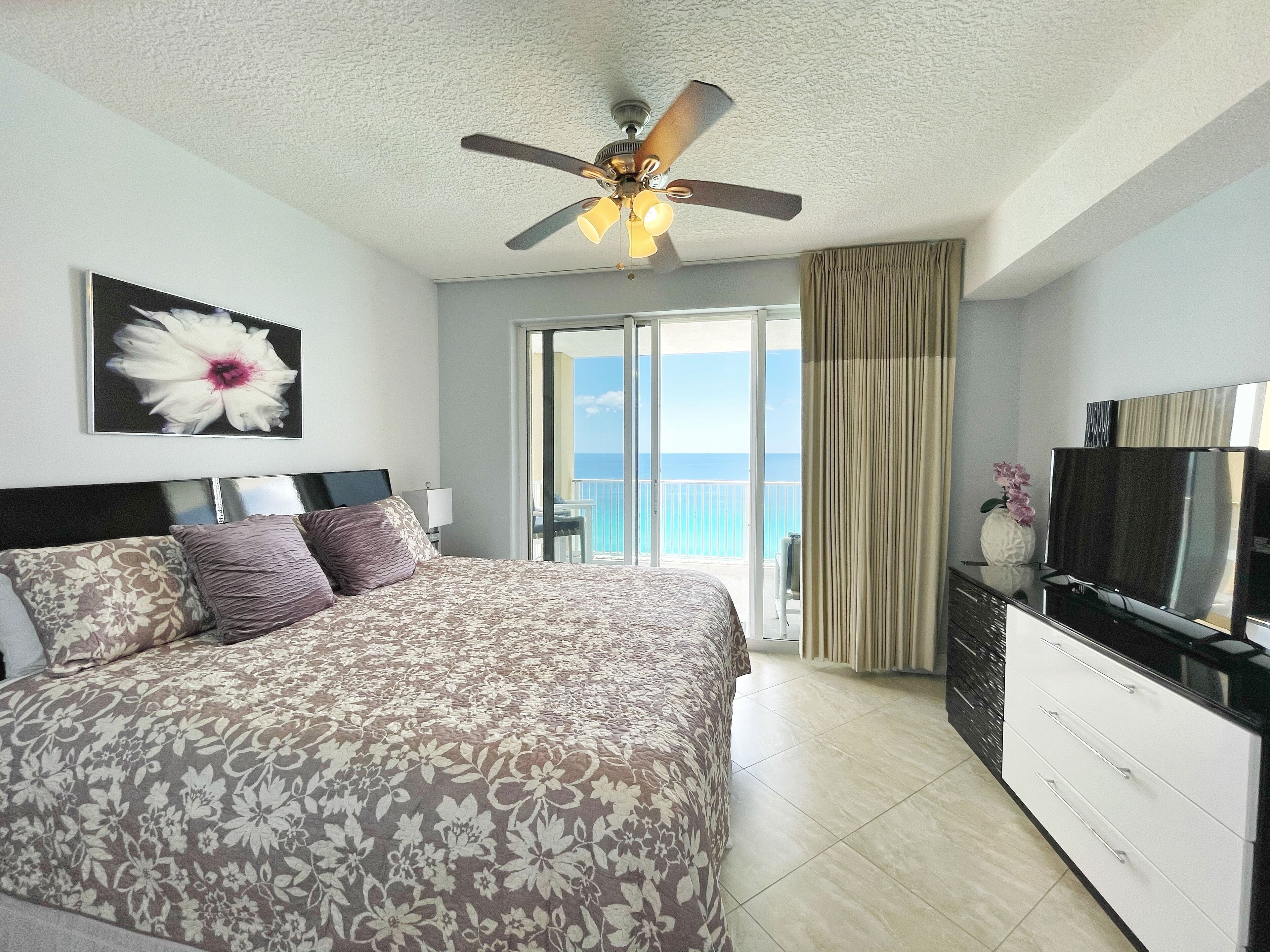 A hotel room with a bed, a ceiling fan, a TV, a balcony with a view of the ocean, and a flower painting on the wall.
