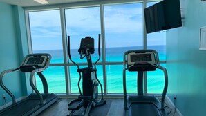 Gimnasio con cintas de correr, bicicleta estática y máquina de remo, con vistas al mar turquesa.