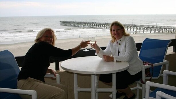 Two people toasting at a round table with a view of the beach and pier in the background. - Serene Oceanfront Getaway Cherry Grove (North Myrtle Beach)3BR (North Myrtle Beach)