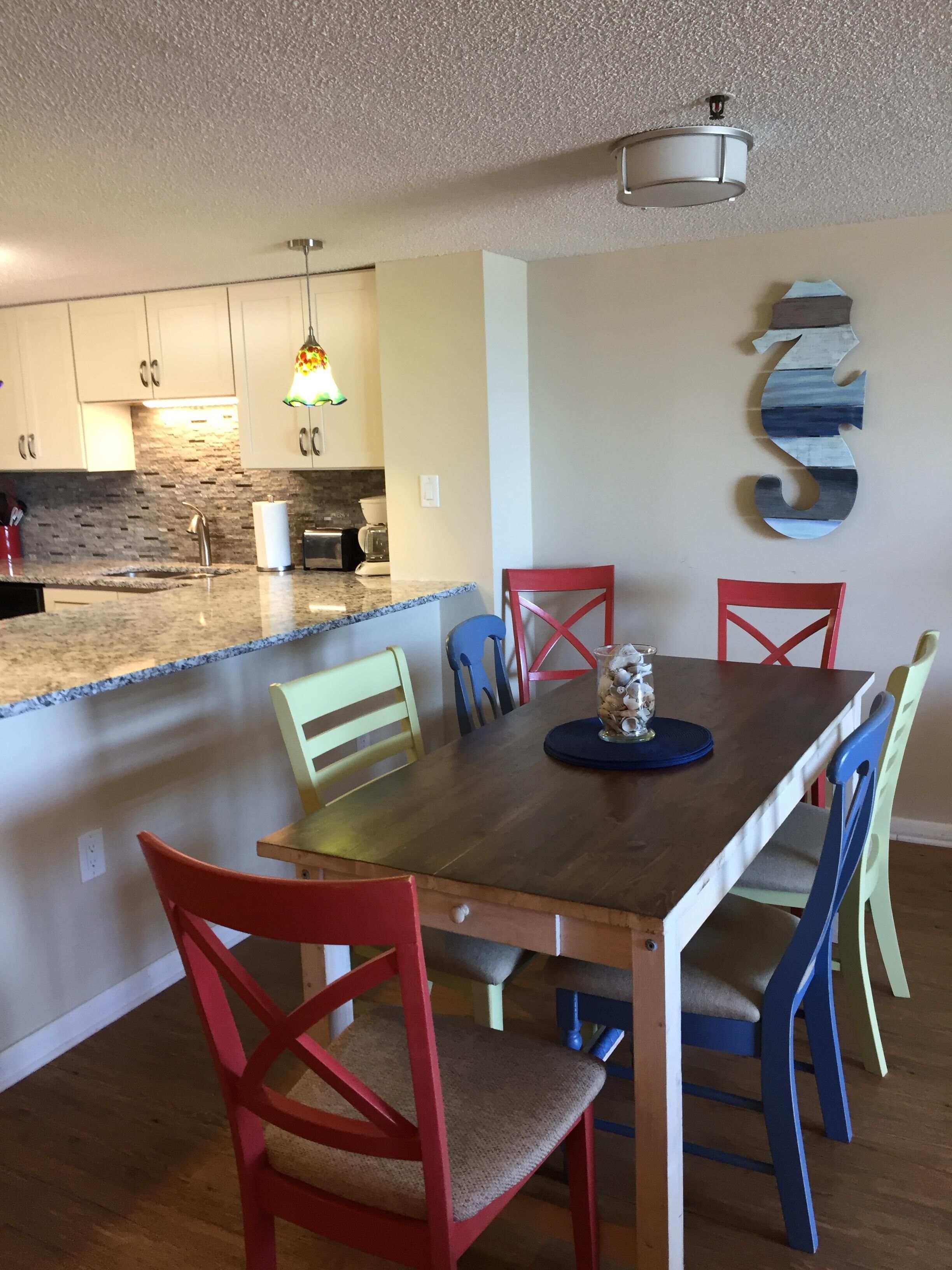 Un coin repas avec une table en bois et des chaises colorées, une cuisine dotée de plans de travail en granit et un élément décoratif mural.