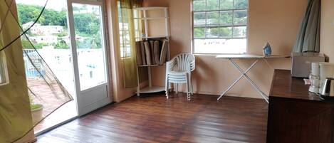 A room with wooden flooring, a white chair, a small table, and a window with a view of greenery and buildings.