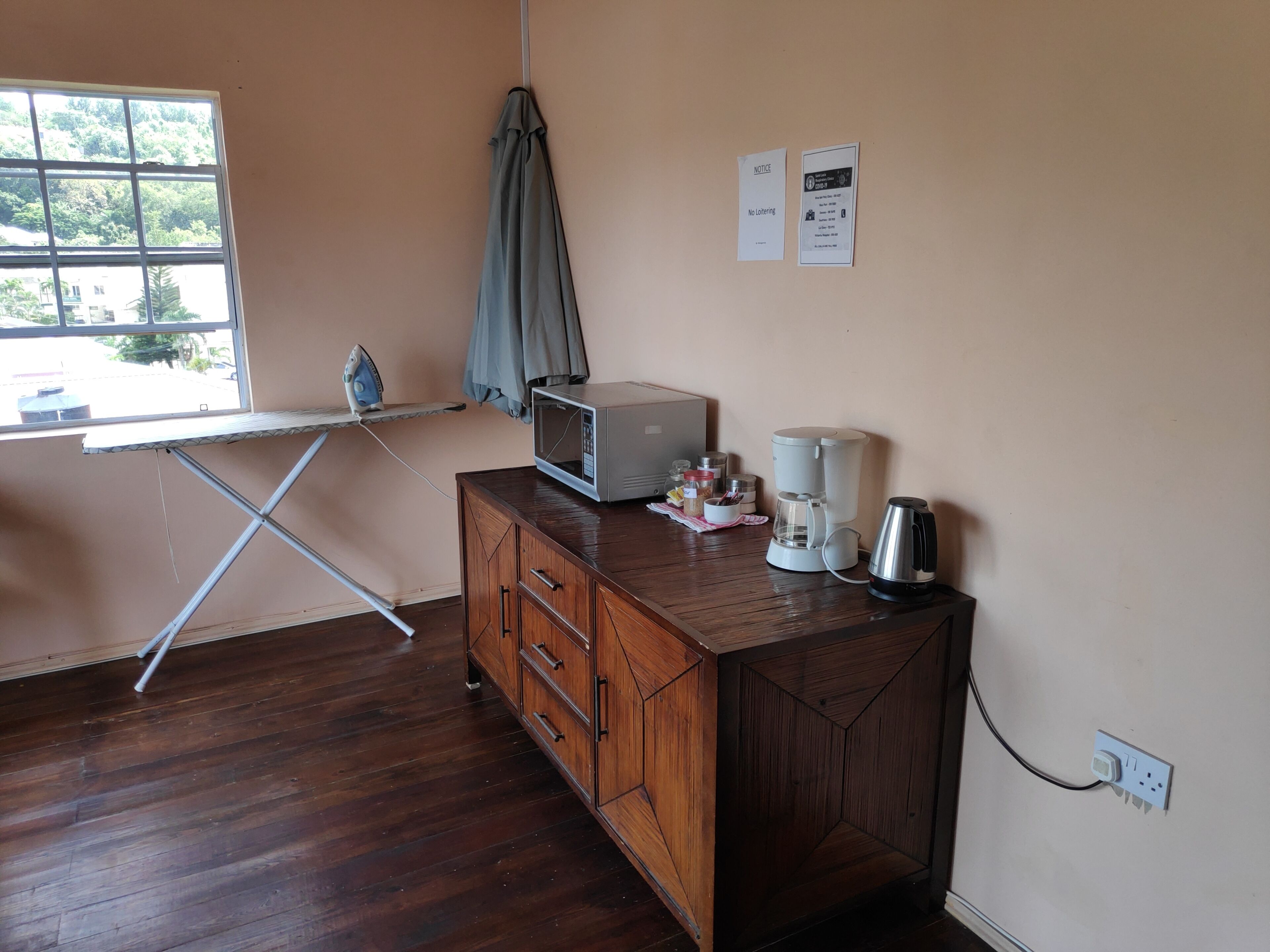 A room with a wooden cabinet, an ironing board, a microwave, a coffee maker, and a window with a view of greenery.