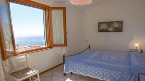 A bedroom with a bed, a chair, a window with a view, and a wall-mounted picture. - VILLETTA LA TERRAZZA SU CHIESSI (Marciana)
