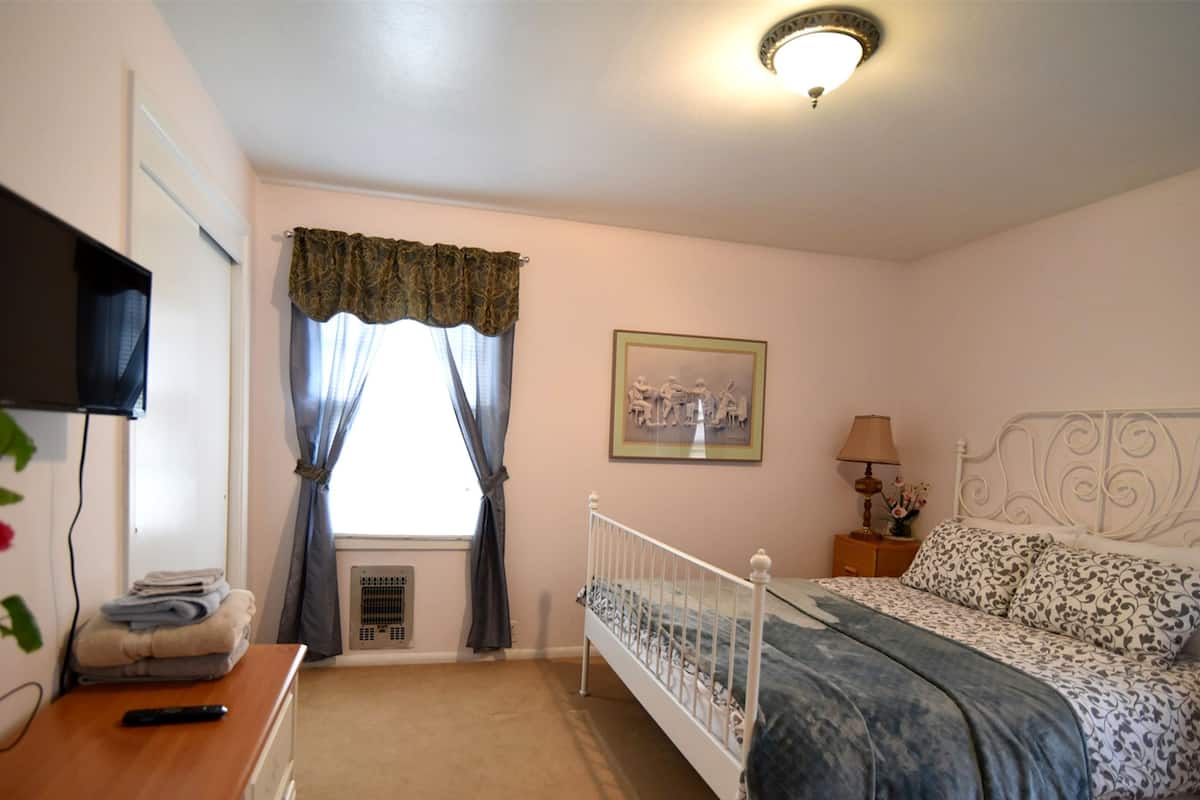 A bedroom with a bed, a bedside table with a lamp, a TV, and a window with curtains.