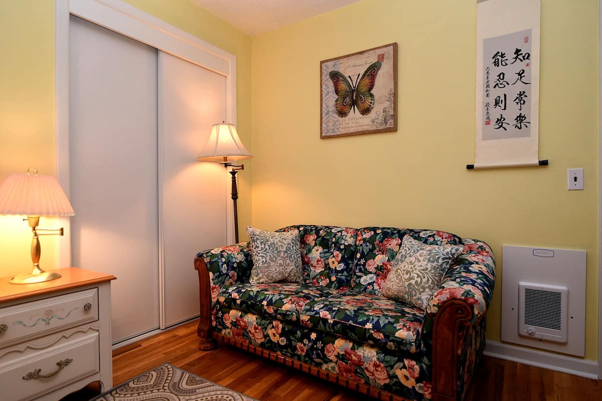 A room with a floral sofa, a lamp, a framed butterfly artwork, and a wall-mounted air conditioner.