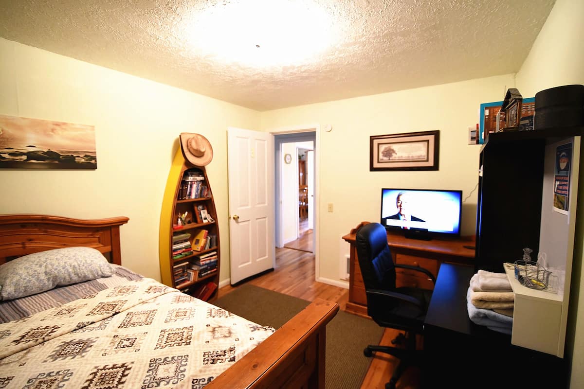 A bedroom with a bed, a desk with a computer, a bookshelf, and a door leading to another room.