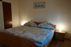 Ein ordentlich bezogenes Bett mit einem gemusterten Überwurf und Kissen, ein Nachttisch aus Holz mit einer Lampe und einer kleinen Schublade.