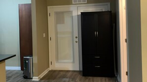 Quarto com piso de madeira, porta branca, armário escuro e lixeira.