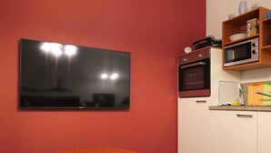 Ein kompakter Wohnbereich mit Flachbildfernseher an der Wand, einer Küchenzeile inklusive Mikrowelle und einem kleinen Holztisch mit Fernbedienung.