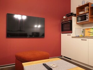 A compact living space with a flat-screen TV mounted on the wall, a kitchenette with a microwave, and a small wooden table with a remote control.