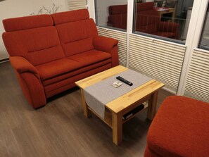 A red sofa, a wooden coffee table with a remote and magazine, and a window with blinds.