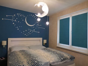 A bedroom with a bed, a wall mural of stars and a crescent moon, a window with blinds, and a nightlight.