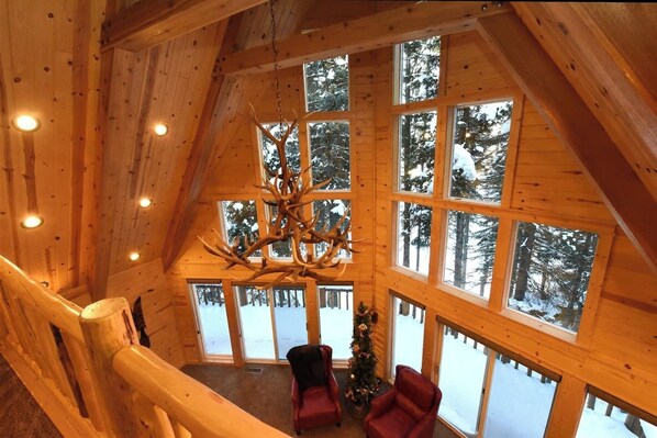 Una cabaña de troncos con un ventanal grande, un candelabro de cornamentas de ciervo y un árbol de Navidad.
