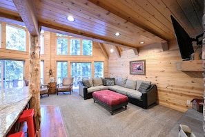 Interior acogedor de una cabaña con un techo de madera grande, un sofá modular, un reposapiés rojo y un comedor con mesa de madera y sillas rojas.