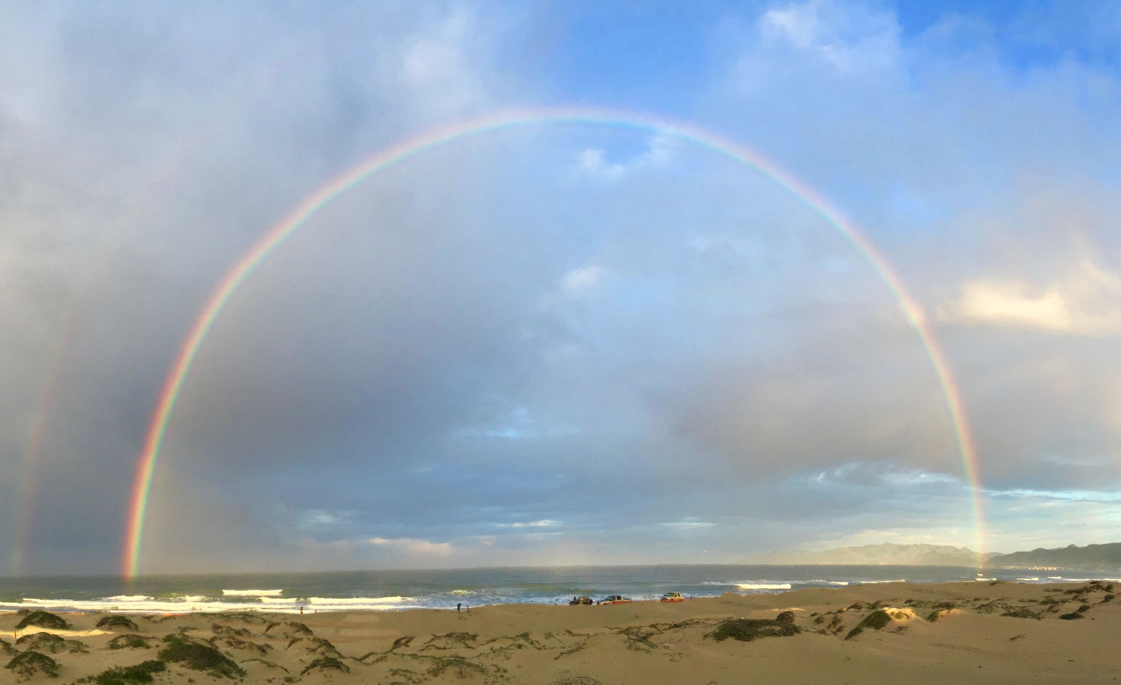 Una playa con un arcoíris doble, dunas de arena y el océano al fondo.