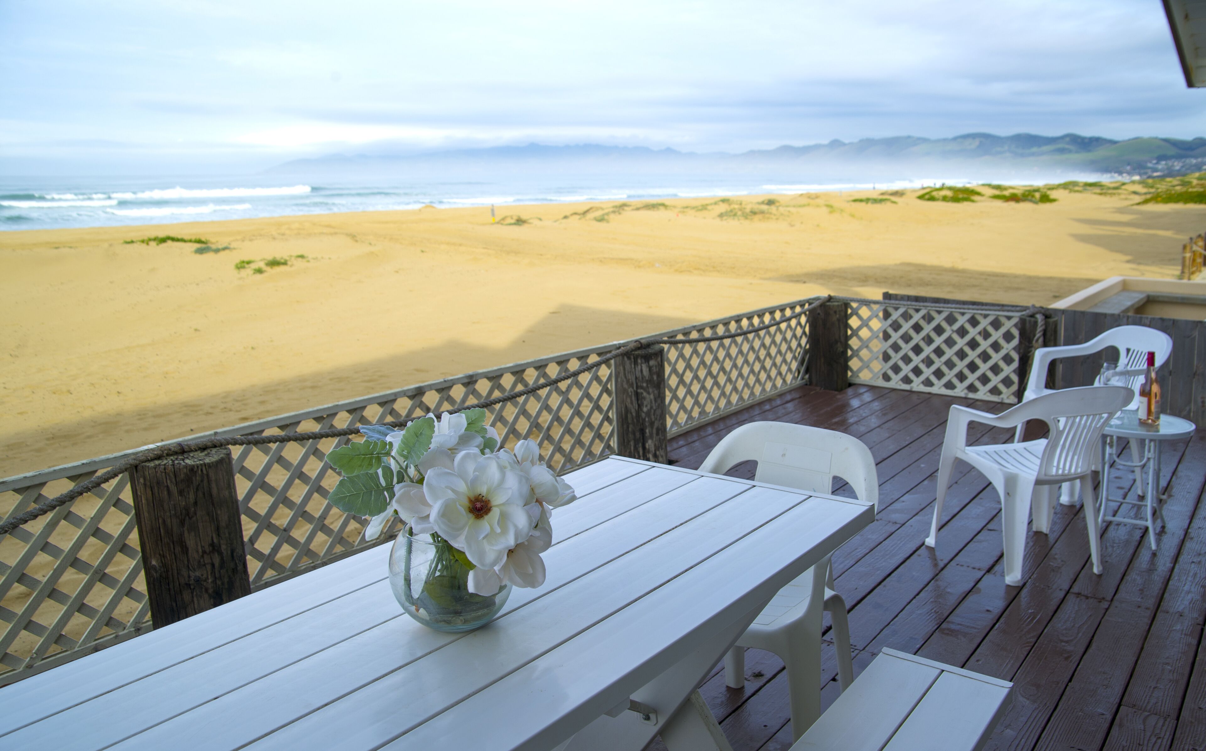 A wooden deck with a table and chairs, overlooking a beach with sand dunes and the ocean.
