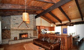A cozy living room with a stone fireplace, leather sofa, wooden ceiling beams, and a kitchen area in the background.