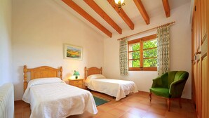 Une chambre avec deux lits simples, un fauteuil vert, une fenêtre avec des rideaux à motifs et un tableau accroché au mur.
