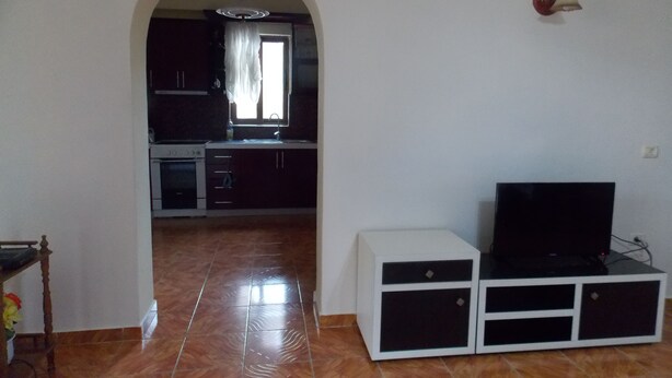 Amplia sala de estar con piso de baldosas, un televisor sobre un mueble blanco y una cocina visible a través de un arco.