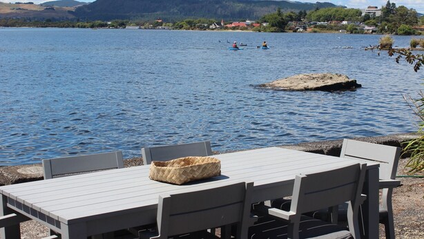 A lakeside table with chairs and a woven basket, overlooking calm water with kayakers in the distance. - Lakeside apartment with boat ramp & jetties (Koutu)