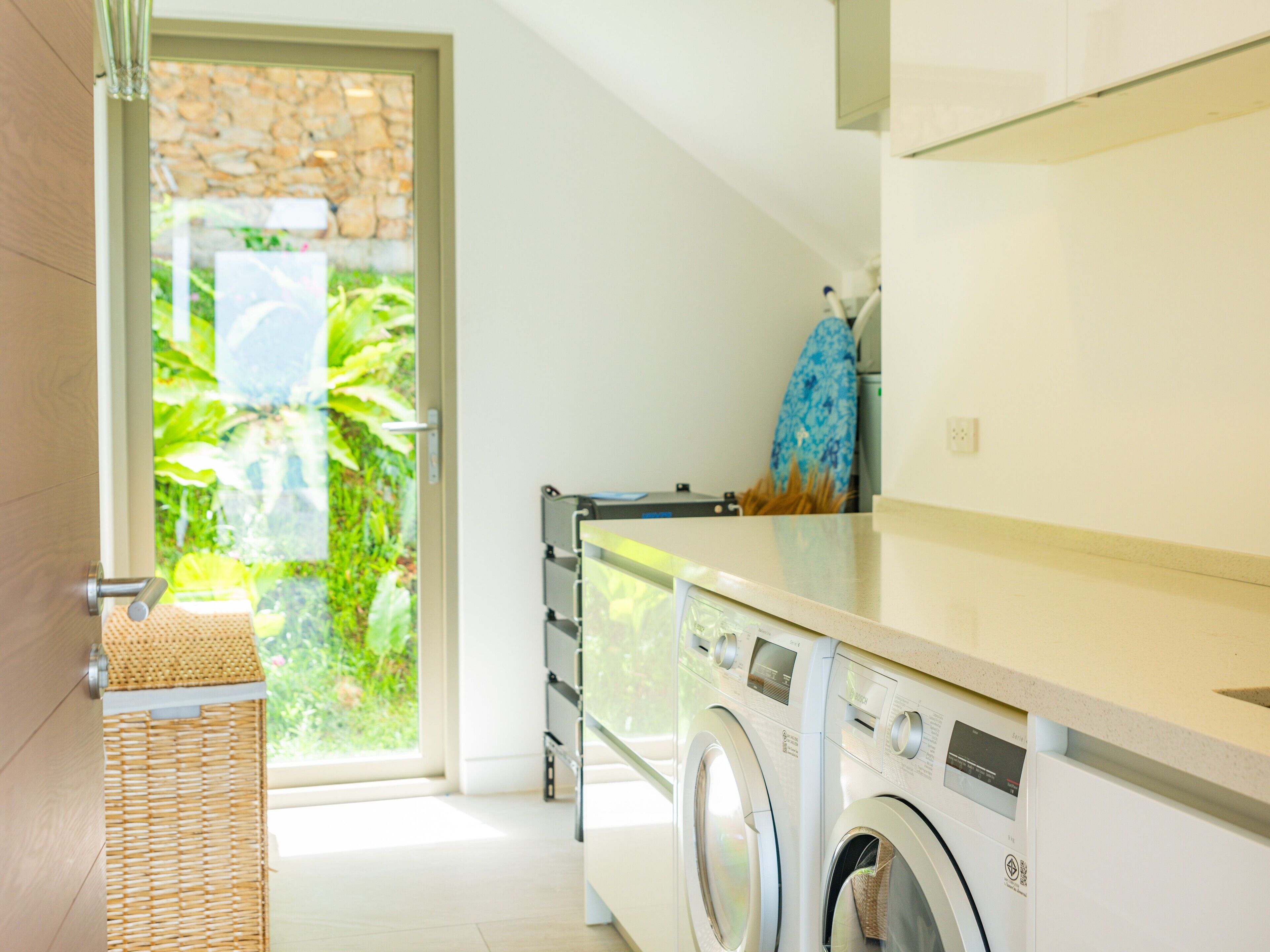 A laundry area with a washing machine and dryer, a wicker basket, and a view of greenery through a glass door.