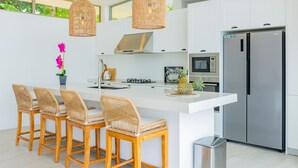 A modern kitchen with a central island, bar stools, a microwave, a stainless steel refrigerator, and wicker pendant lights.