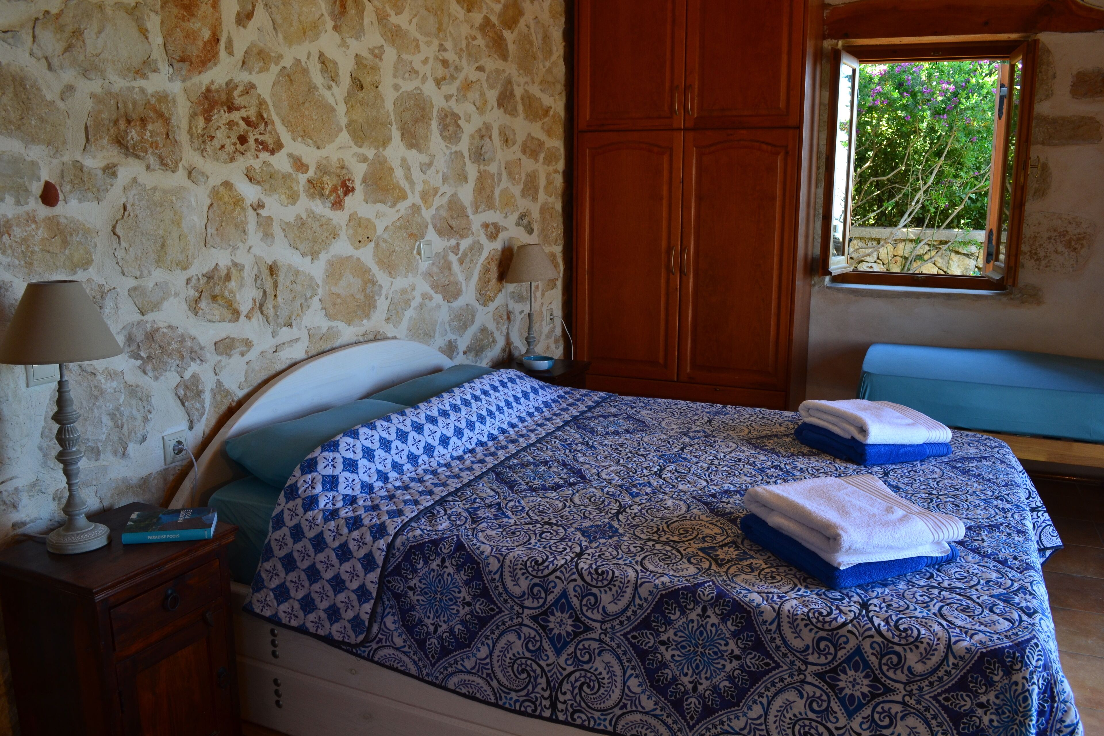 A bedroom with a stone wall, wooden wardrobe, and a window with a view of greenery.