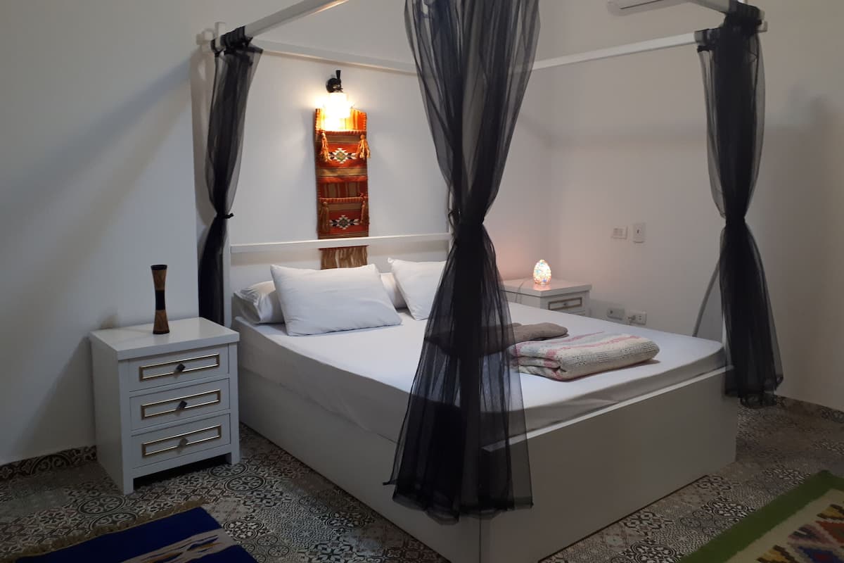 A bedroom with a canopy bed, a bedside table with a lamp, a decorative wall hanging, and a patterned rug.