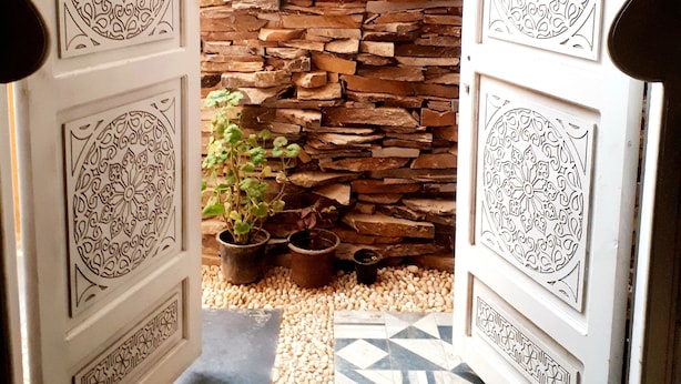 An open doorway with intricate white designs, leading to a stone wall and a small garden area with potted plants. - Arabian Nights Pyramids Guesthouse (Giza)