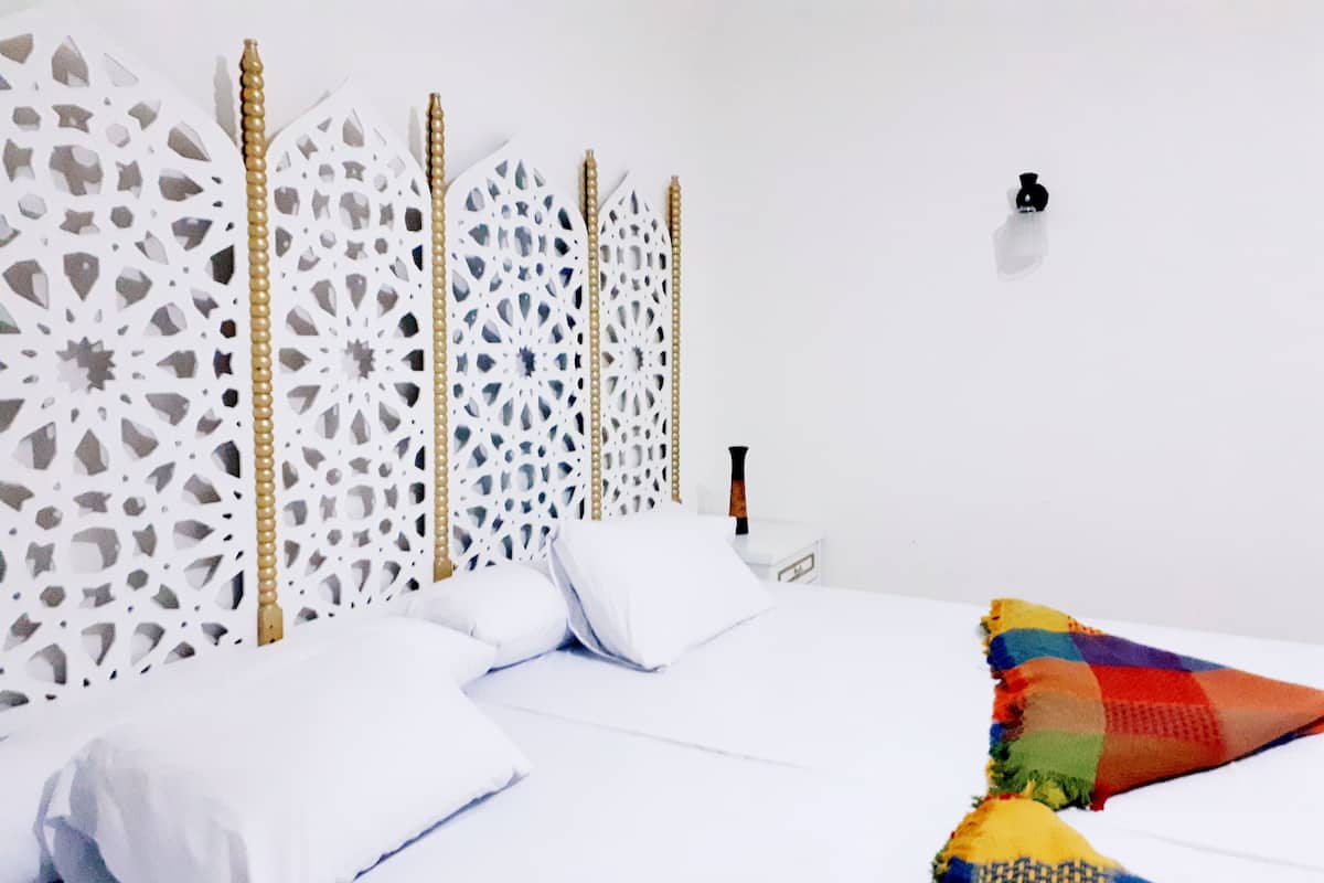 A neatly made bed with white linens and a colorful blanket, a wooden headboard with intricate cut-out patterns, and a small bedside table with a bottle.