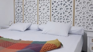 A bed with white linens and a colorful blanket, a white nightstand with a decorative item, and a wall with intricate white panels. - Arabian Nights Pyramids Guesthouse (Giza)