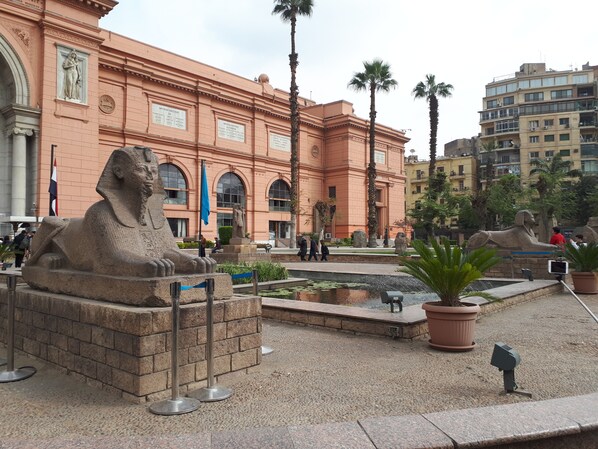 Une grande statue de sphinx se dresse devant un bâtiment doté d’arcades et surmonté d’un drapeau.
