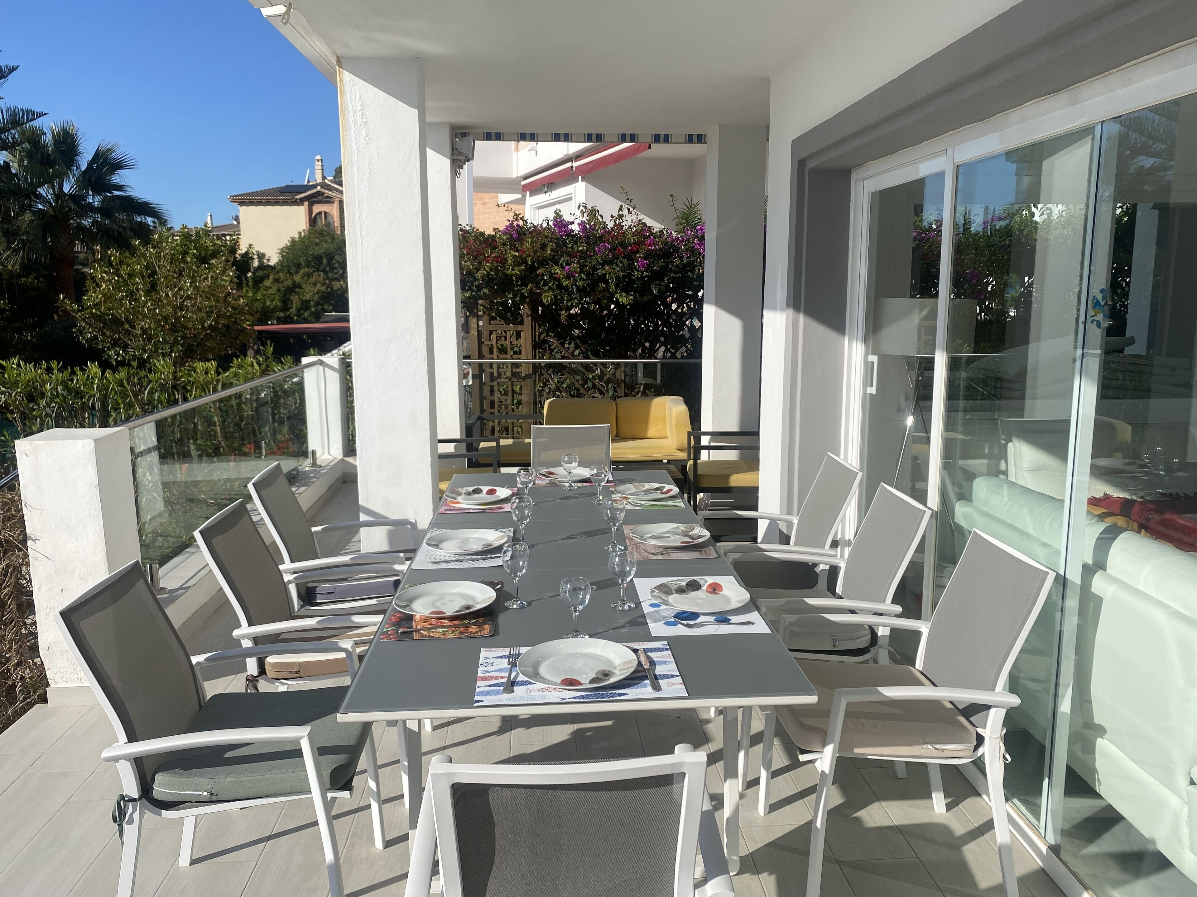 Un área de comedor al aire libre con una mesa dispuesta para seis personas, rodeada de sillas blancas.