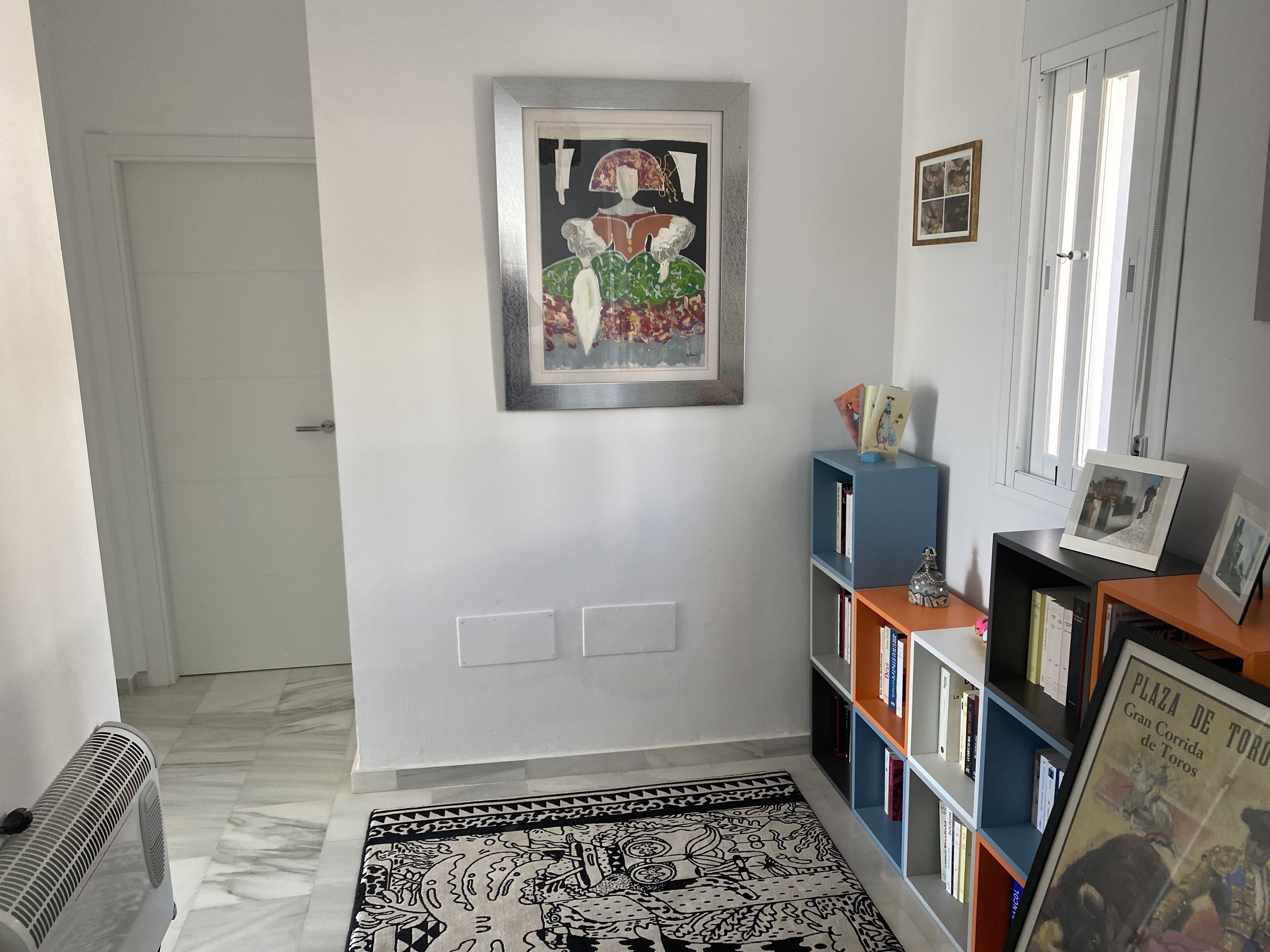A hallway with a patterned rug, a framed artwork on the wall, and a bookshelf with books and decorative items.