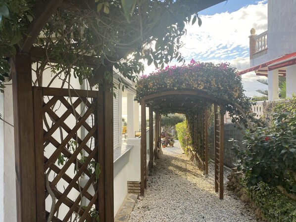 A covered walkway with a wooden lattice gate, gravel path, and a pergola with hanging flowers. - villa halcyon 24 calle huelva calahonda (Mijas)