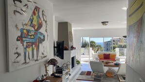 A modern living room with a large abstract painting, a flat-screen TV, and a dining area with a white sofa and colorful cushions. - villa halcyon 24 calle huelva calahonda (Mijas)
