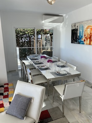 Un espace de salle à manger moderne, avec une longue table dressée pour six personnes, des chaises blanches et une vue sur un jardin, visible à travers de grandes fenêtres.