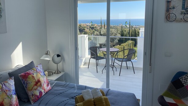 A bedroom with a bed, a nightstand, a lamp, and a view of the balcony with a table and chairs. - villa halcyon 24 calle huelva calahonda (Mijas)
