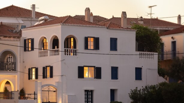 A waterfront building with multiple balconies, a boat docked in front, and a calm water body reflecting the building. - Porto Vecchio Luxury Suites (Spetses)