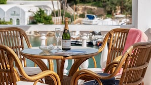 A wicker table with a bottle of champagne, a glass, and a menu on a balcony overlooking a marina with boats. - Porto Vecchio Luxury Suites (Spetses)