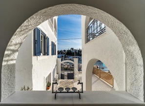 Une vue à travers une fenêtre en arc d’un bâtiment à la façade blanche, aux volets bleus, donnant sur un port où se trouvent des bateaux.
