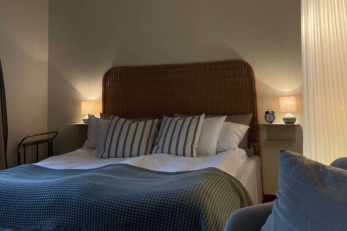 Een keurig opgemaakt bed met een gestreept kussen, een bedlampje en een wandklok op een nachtkastje.