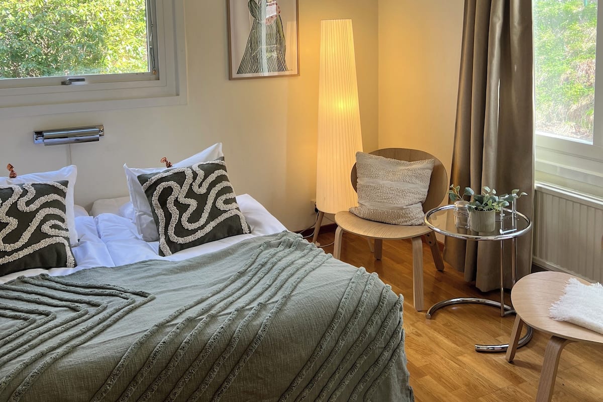 Een slaapkamer met een bed, een stoel, een tafel met een plant en een raam met gordijnen.