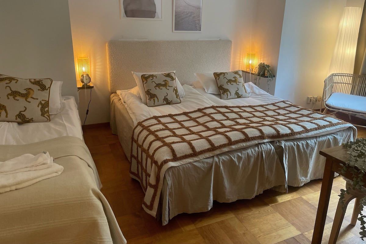 Een hotelkamer met twee bedden, een groot bed met een bekleder met patroon en een kleiner bed. Er zijn wandlampen en een klok op het nachtkastje.